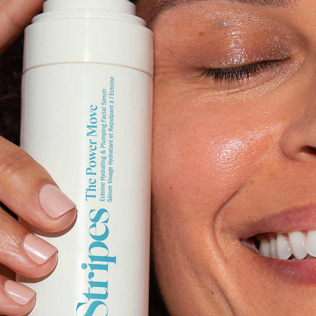 Close-up of a smiling woman holding Stripes The Power Move hydrating and plumping facial serum bottle.
