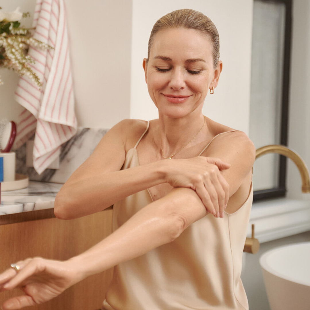 Naomi Watts applying oil on her arm in a bathroom.