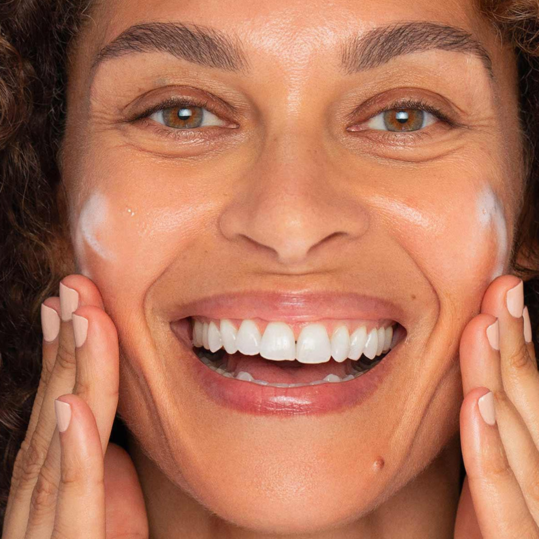 Close-up of a smiling woman applying Resting Clean Face creamy cleanser on her cheeks for hydration and soothing.