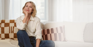 Naomi Watts sitting on a couch with patterned cushions in a bright room