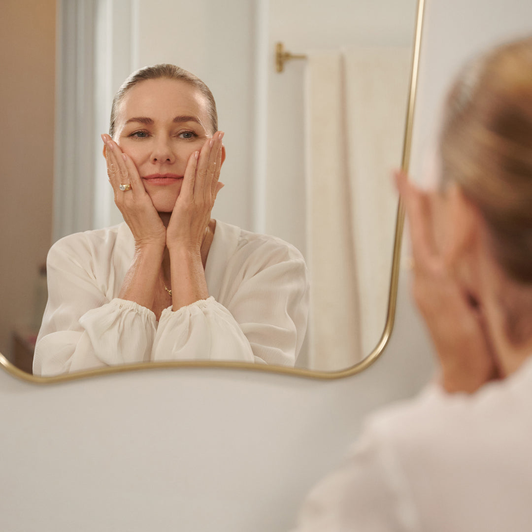 Naomi Watts in a white robe looking at herself in a mirror, touching her face.