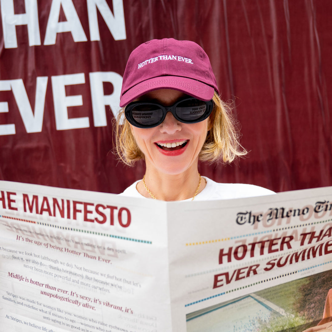 Naomi Watts wearing a pink cap and sunglasses, holding a newspaper with 'HOTTER THAN EVER SUMMER' headline. She is wearing the Hotter than July Stripes Beauty Hat.