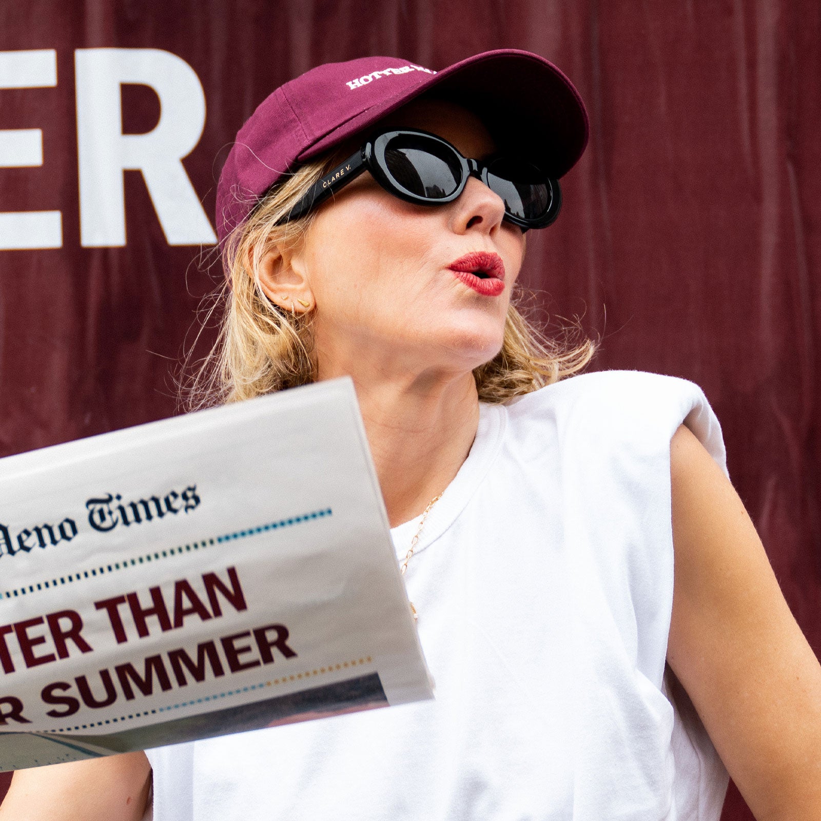 Naomi Watts wearing sunglasses and a cap, holding a newspaper with text, against a red background. She is wearing the Hotter than July Stripes Beauty Hat.