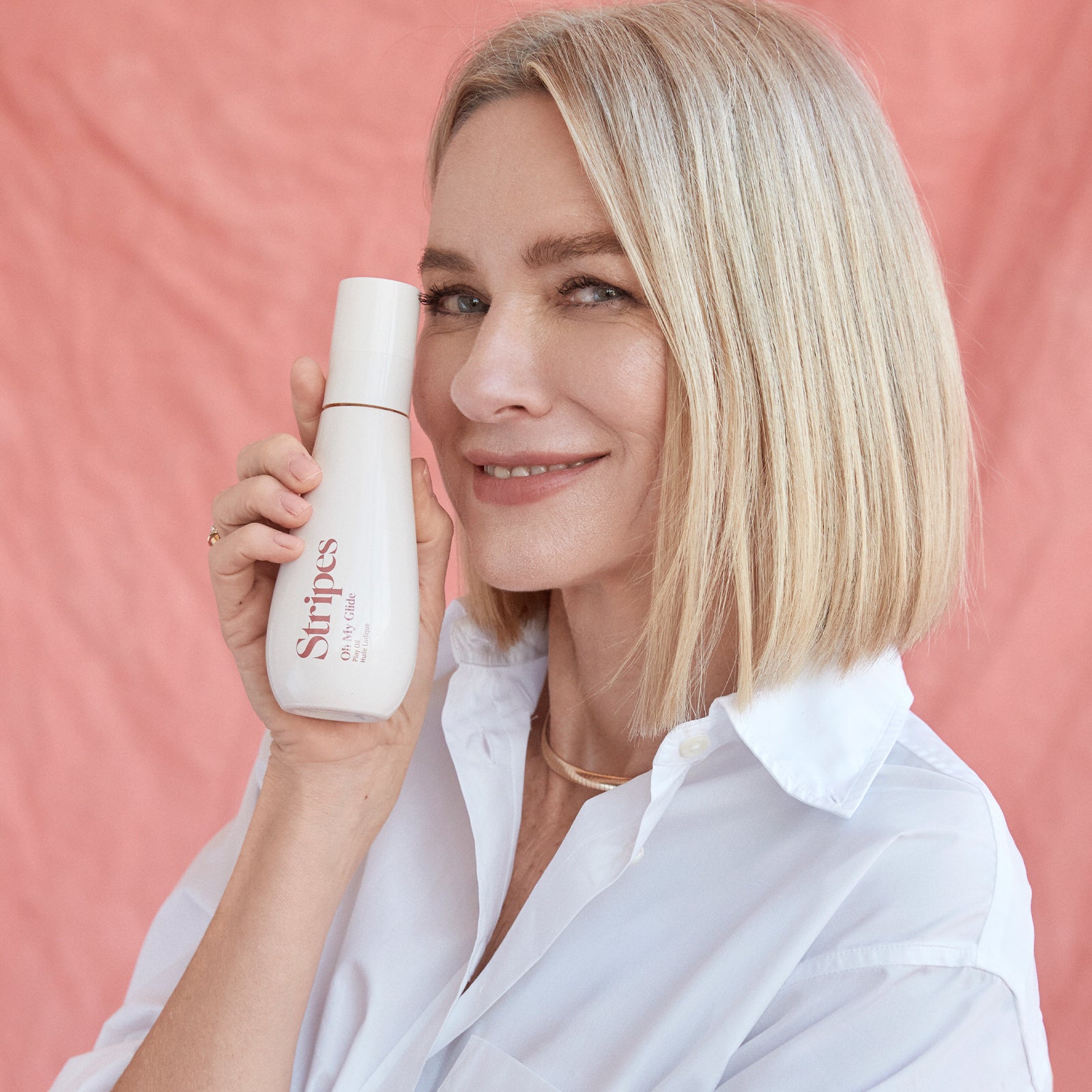 Stripes Beauty Vag of Honor bottle held by smiling Naomi Watts a soft pink background.